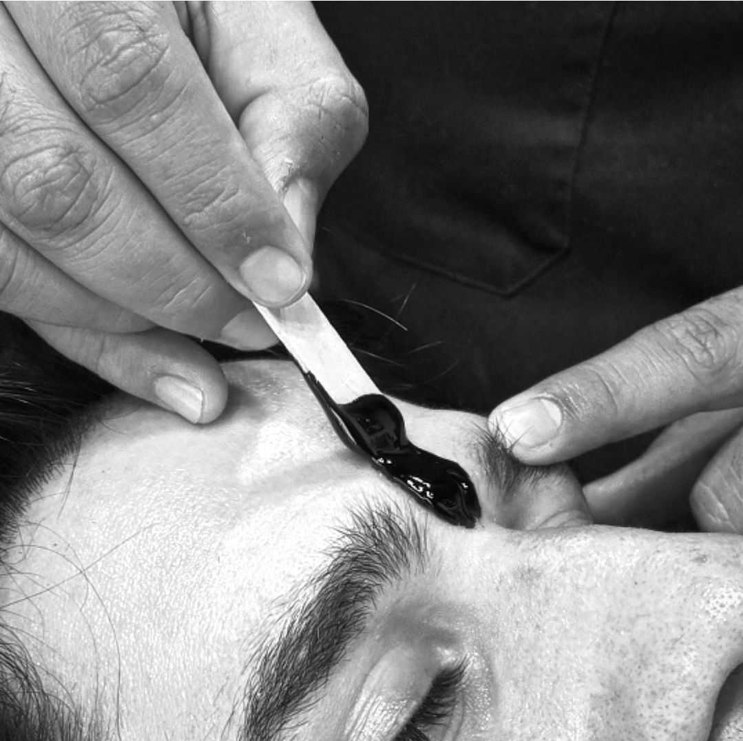 Applying wax to shape a man's eyebrows with a wooden stick in a close-up view.
